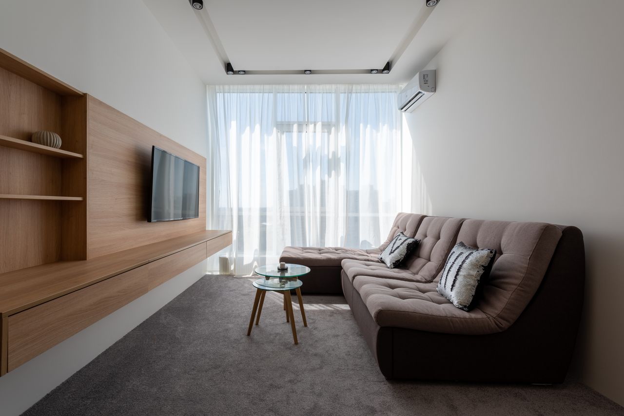 Comfortable couch with cushions near side tables against wall with wooden cupboard and modern TV in stylish living room with window